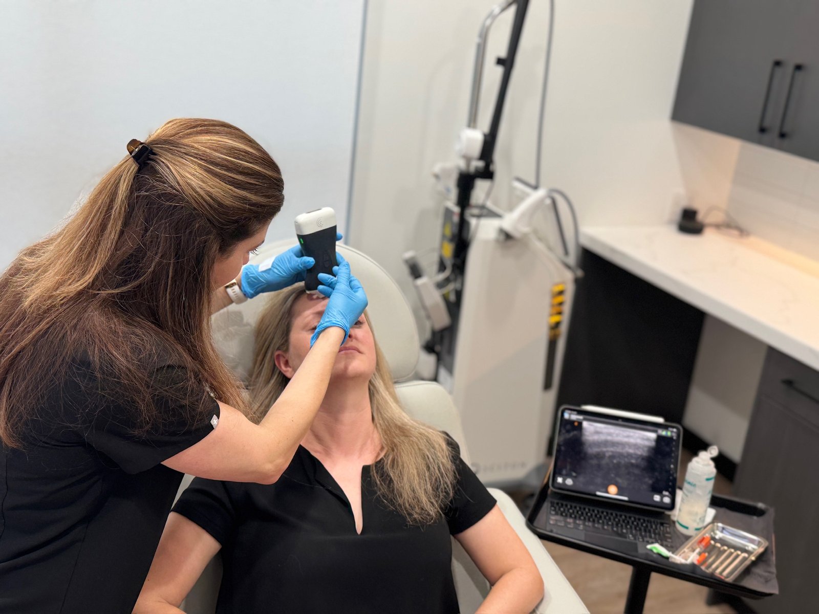 Laura Osterweil performing an ultrasound-guided facial injection on a female patient, holding a probe near the forehead area.